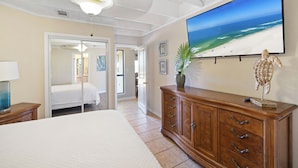 Quarto com uma cama grande, cômoda de madeira, televisão exibindo uma cena de praia e porta que dá para outro espaço.