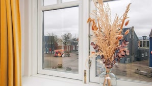 Ein Fenster mit einer Vase gefüllt mit getrockneten Pflanzen und Blick auf einen Stadtplatz mit Gebäuden.