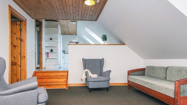 Ein Loft-Zimmer mit schrägem Dach, einem grauen Sessel, einer Holzbank und einem kleinen Tisch.