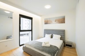 A modern bedroom with a large bed, a small bedside table, a nightstand, and a window with a view of buildings. - Modern Lisbon Balcony AND Pool by Homing (Lisbon)