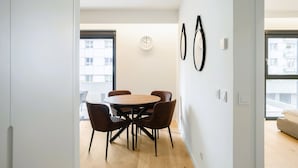 Uma área de refeições moderna com uma mesa redonda e quatro cadeiras, um relógio na parede e uma janela com vista para edifícios.