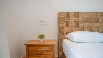Une chambre à coucher avec une table de chevet en bois, une petite plante en pot, un lit avec une literie blanche et une tête de lit en osier.