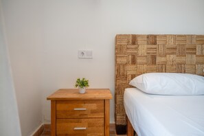 Ein Schlafzimmer mit einem Nachttisch aus Holz, einer kleinen Topfpflanze, einem Bett mit weißer Bettwäsche und einem gewobenen Kopfteil.
