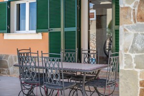 An outdoor seating area with metal chairs and a table, a window with green shutters, and a stone wall. - Dream Villa with Private Pool in Cala Romántica (S'Estany d'en Mas) (Romàntica)