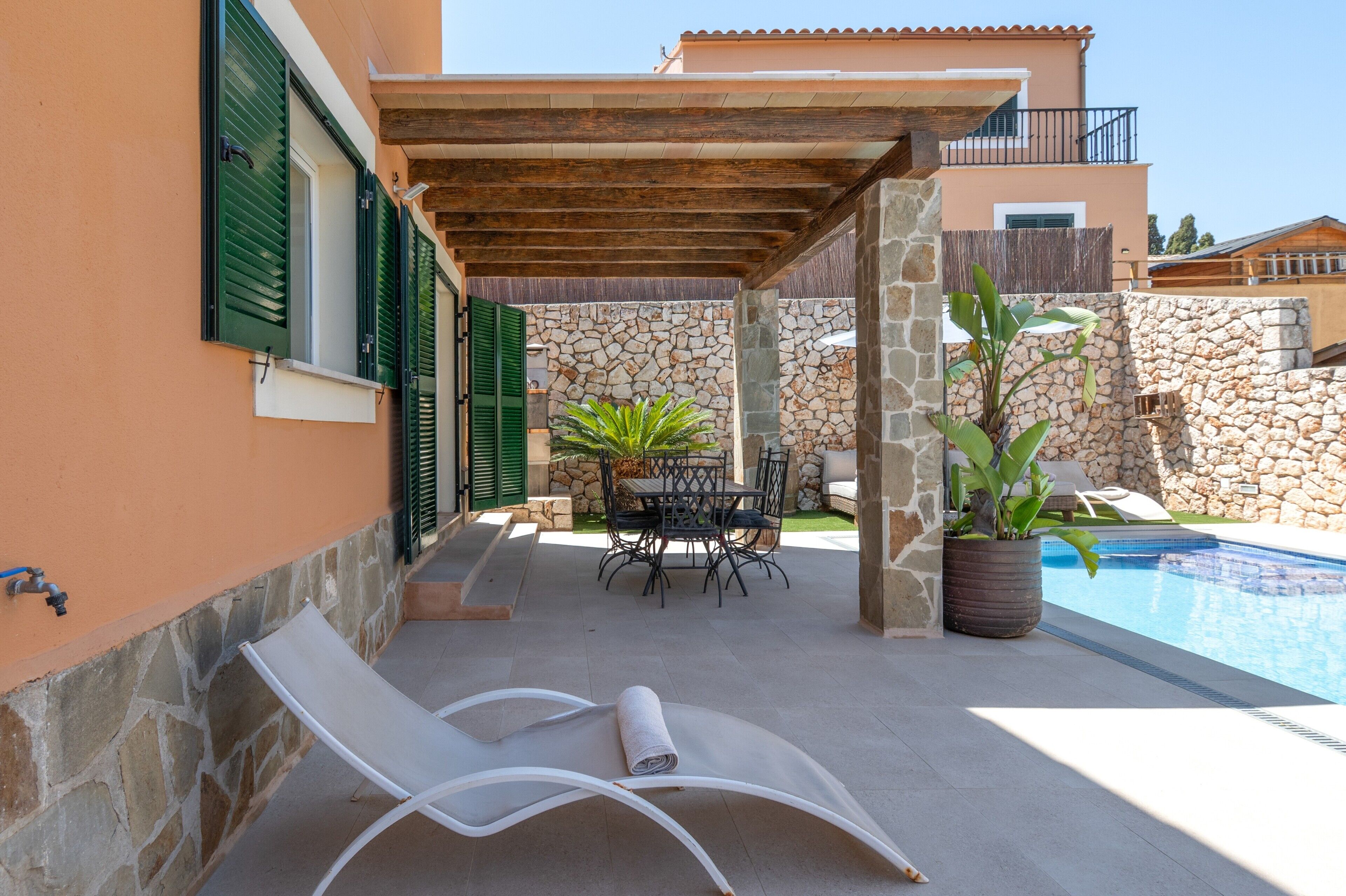 Un área junto a la piscina con una pérgola, una mesa con sillas, una tumbona y una planta en maceta.