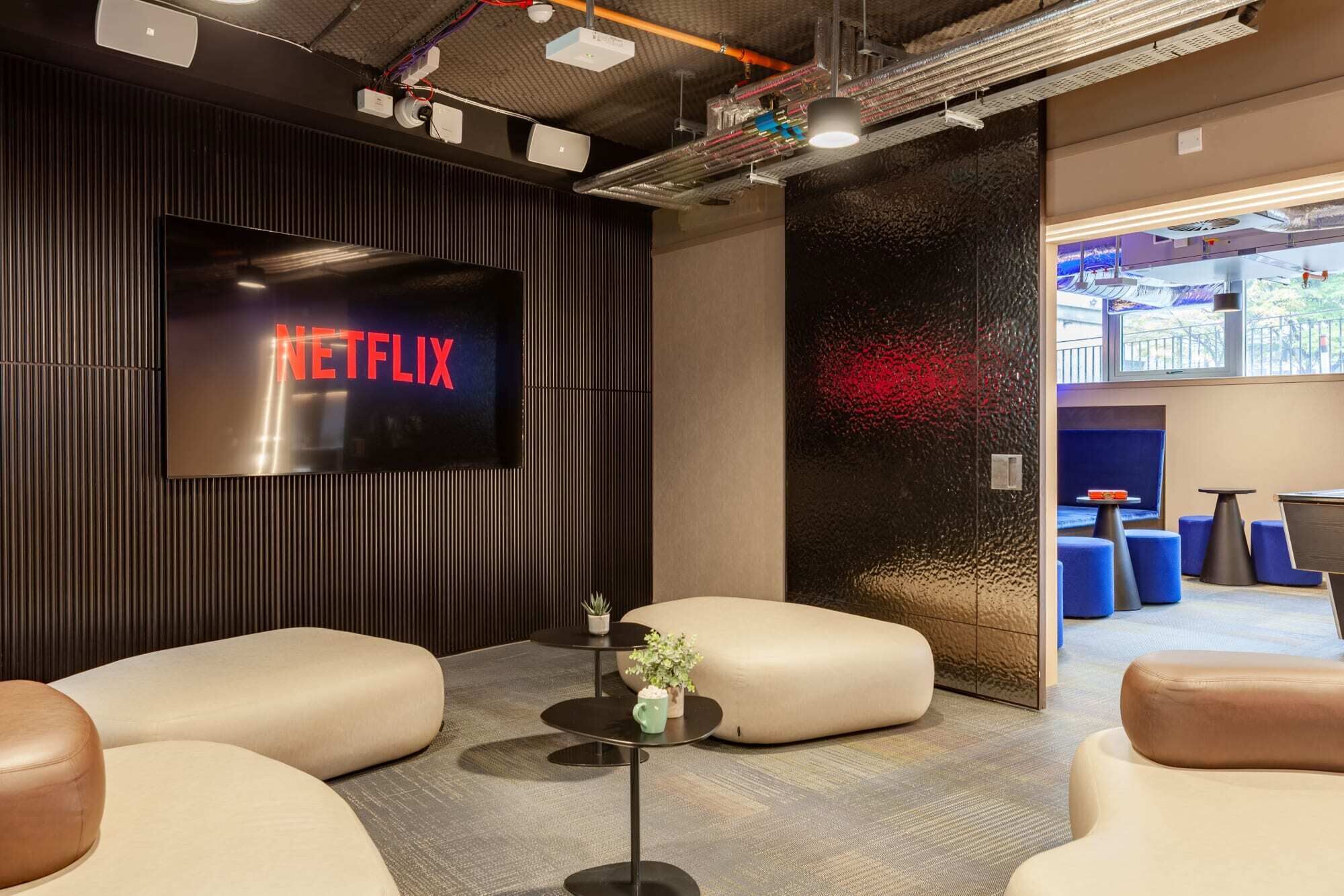 A modern lounge area with a large flat-screen TV displaying the Netflix logo, comfortable seating, and a view of the outdoors through a large window.
