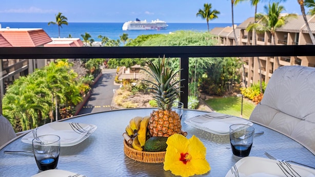 A dining table with a tropical fruit arrangement, a hibiscus flower, and a view of the ocean and cruise ship. - Kona Pacific E734: Penthouse Corner unit, Best Ocean Views, AC, Walk to town (Kailua Kona)