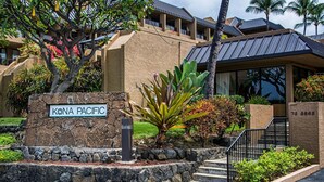 A building with a stone sign reading 'KONA PACIFIC,' a staircase leading to the entrance, and a well-maintained garden with various plants. - Kona Pacific E734: Penthouse Corner unit, Best Ocean Views, AC, Walk to town (Kailua Kona)