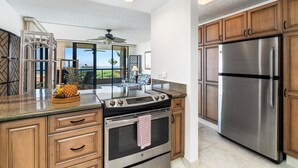 A modern kitchen with wooden cabinets, stainless steel appliances, and a large island with a gas stove. - Kona Pacific E734: Penthouse Corner unit, Best Ocean Views, AC, Walk to town (Kailua Kona)
