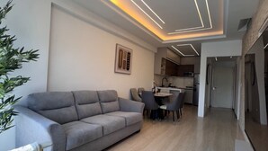Een moderne woonkamer met een grijze bank, een eethoek met stoelen, en een keuken op de achtergrond.