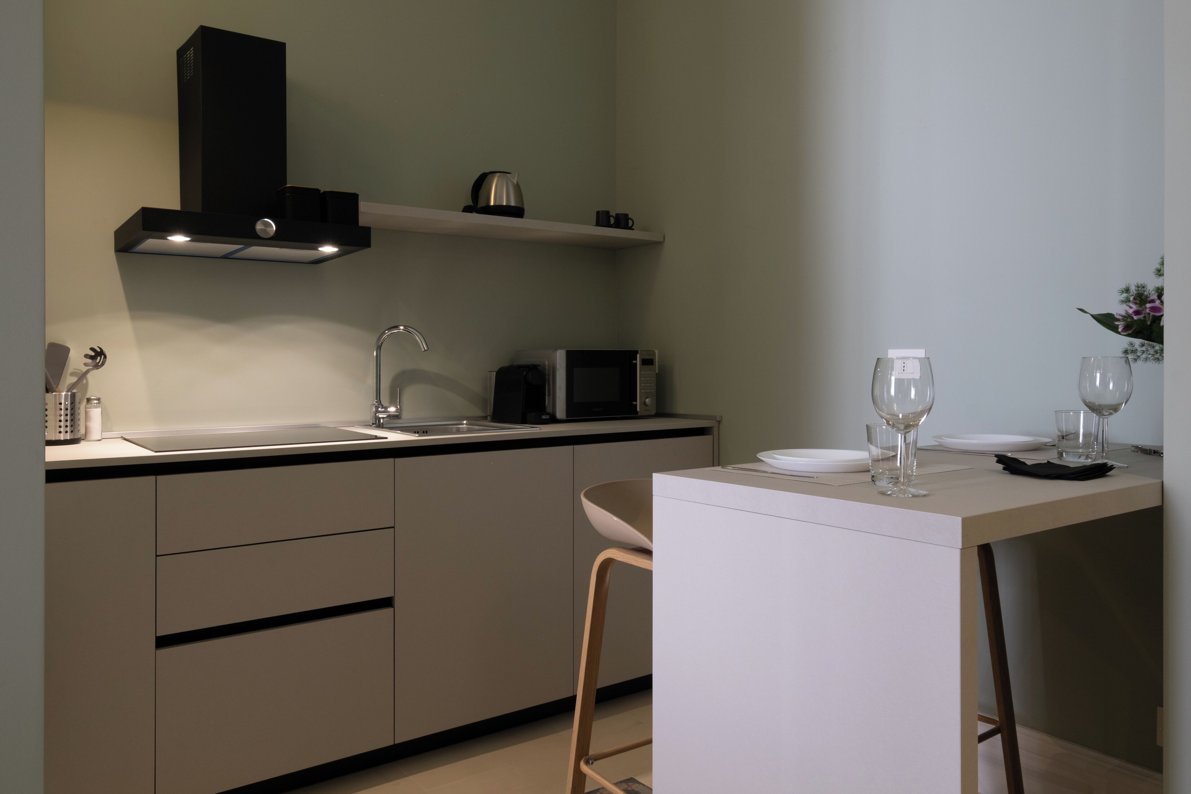 Una cocina moderna con encimera blanca, microondas, hervidor de agua y lavamanos. Hay una zona de bar con una mesa blanca, dos vasos y un plato.