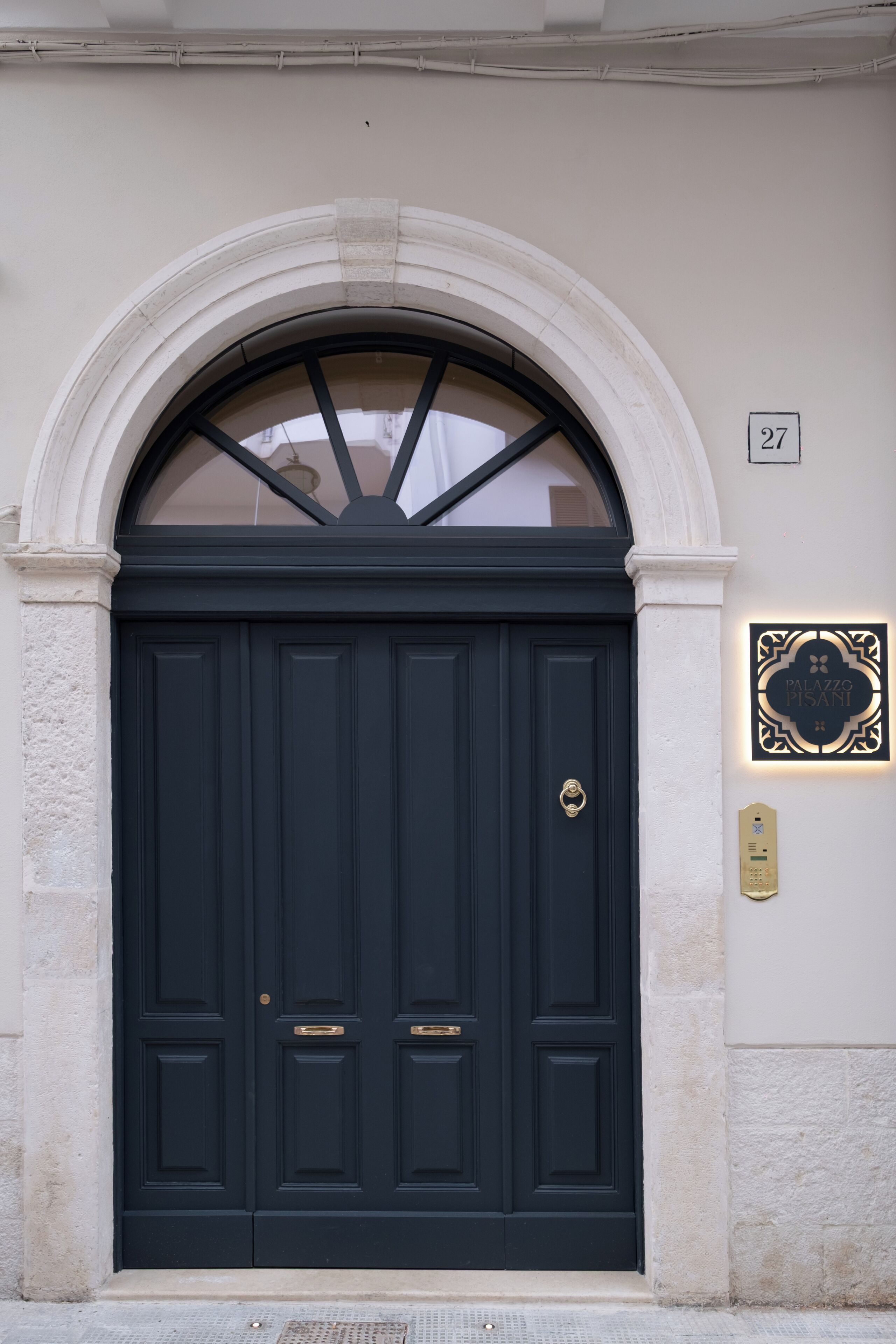 Una puerta azul oscuro con un pomo dorado, enmarcada en un arco de piedra blanca. Un letrero con el número 27 y el nombre "Palazzo Pisani" está colocado en la pared a la derecha.