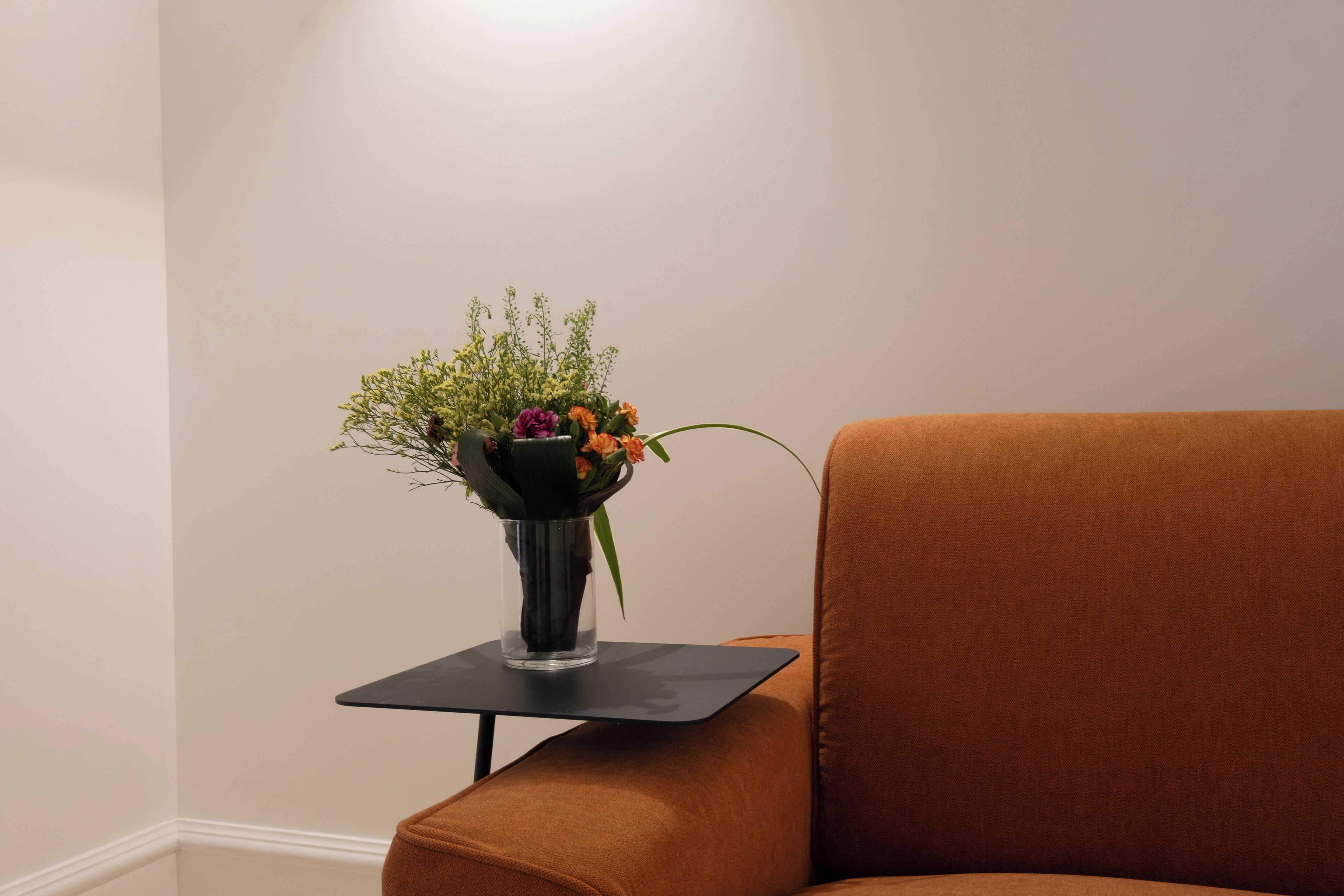 Esquina de una habitación con un sillón marrón y una mesita pequeña con un florero transparente con flores.