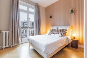 Ein Hotelzimmer mit einem großen Bett, Blick durch ein Fenster, einem Heizkörper und einem Nachttisch mit einer Lampe.