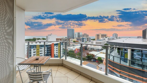 Un balcón con mesa y sillas, con vistas a un paisaje urbano con edificios y un cielo al atardecer.