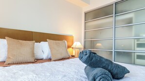 Una cama bien hecha con un edredón blanco, dos almohadas color beige y una toalla gris enrollada.