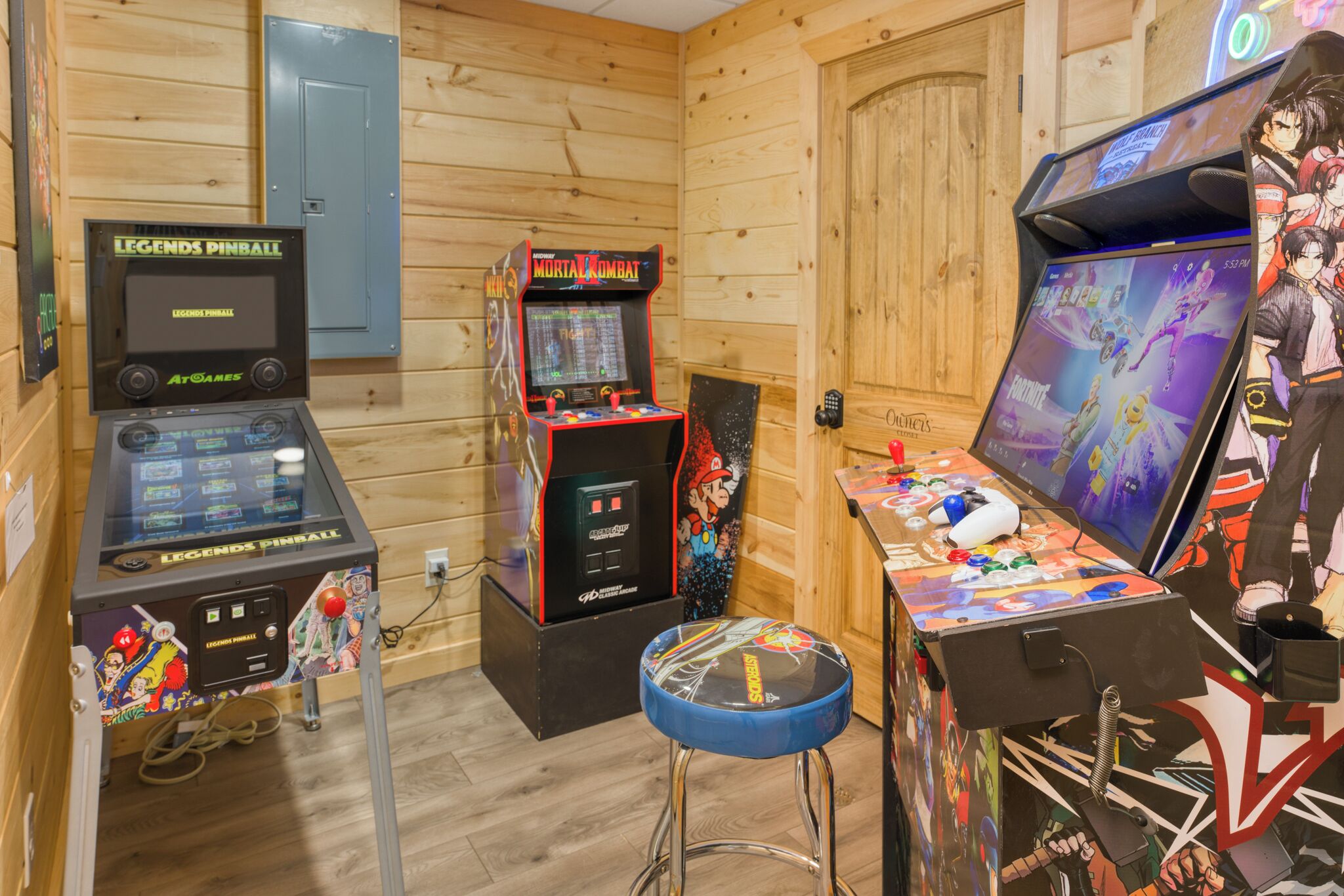 A pinball machine, a Mortal Kombat arcade game, and a Fortnite gaming station in a room with wooden walls and a blue door.