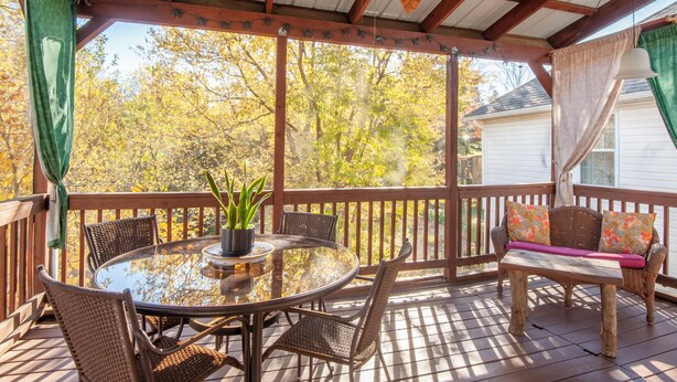 Une terrasse extérieure couverte, dotée d’une table ronde en verre et de chaises en osier, entourée d’une terrasse en bois et offrant une vue sur les arbres.