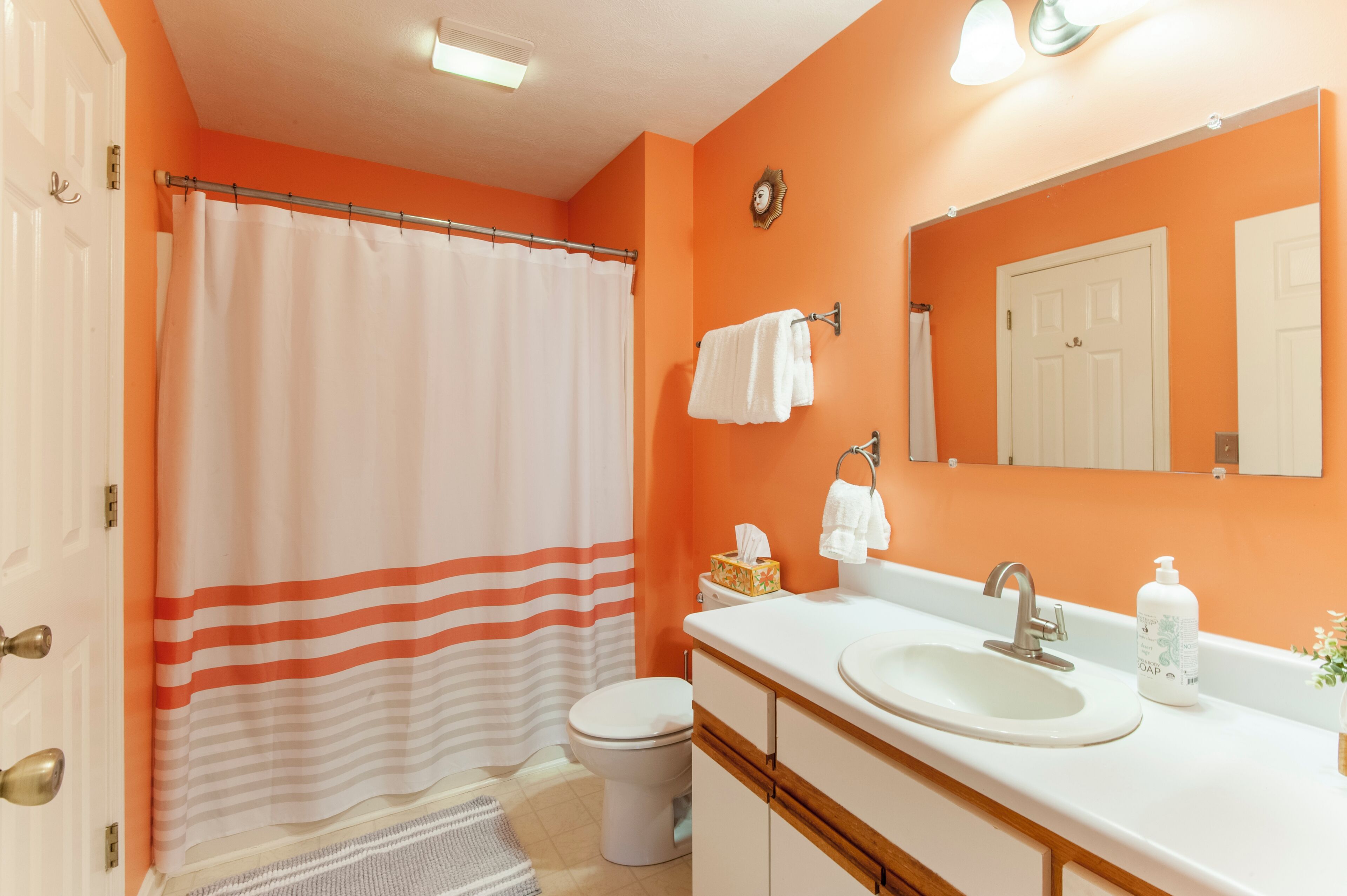 Une salle de bain avec un mur orange, un lavabo blanc, un miroir, des toilettes blanches et un rideau de douche à rayures.