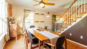 Un espace repas comprenant une longue table en bois, des chaises, un ventilateur de plafond et un escalier menant à un niveau supérieur.