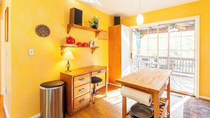 Une pièce lumineuse aux murs jaune vif, avec une table en bois, une poubelle, un bureau avec des tiroirs, une chaise et une fenêtre donnant sur l’extérieur.