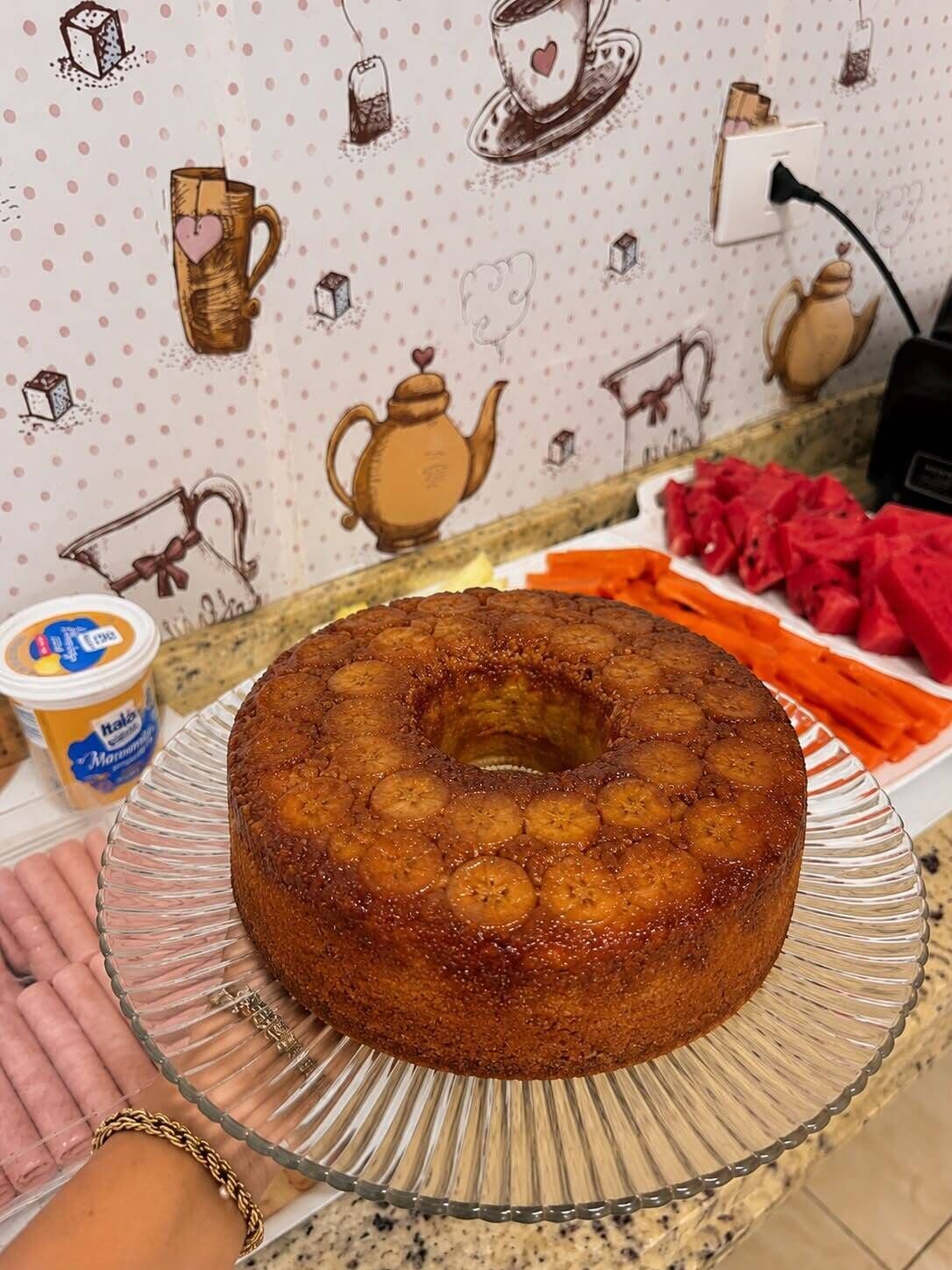 Un gâteau décoré de tranches de banane, présenté sur une assiette en verre, posé sur un plan de travail de cuisine.