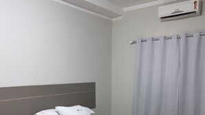 A bedroom with a bed, a bedside table, a window with curtains, and an air conditioning unit. - NOSSO HOTEL (Três Lagoas)