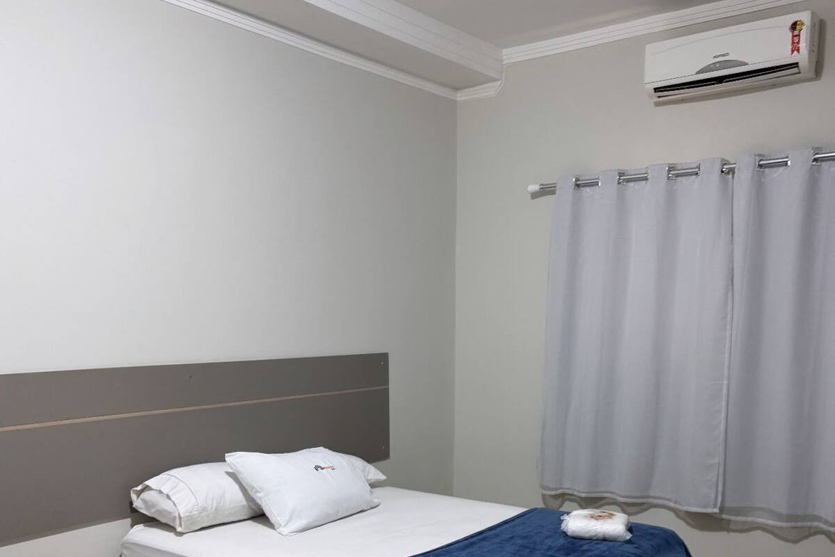 Un dormitorio con cama, mesita de noche, ventana con cortinas y aire acondicionado.