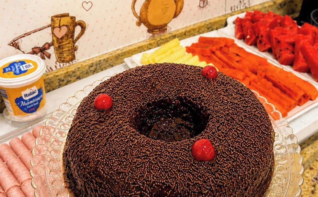 A chocolate bundt cake with sprinkles and cherries on a glass plate, next to a container of yogurt and a plate of sliced fruits.