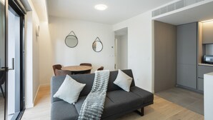 A modern living room with a grey sofa, two white pillows, and a grey throw blanket. There is a dining table with chairs, a kitchen area, and a large mirror on the wall. - Modern Lisbon W Balcony by Homing (Lisbon)