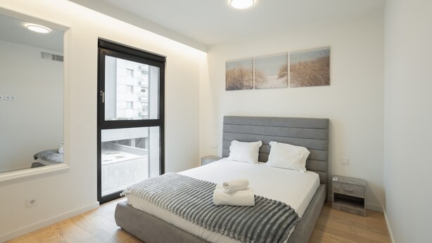 A modern bedroom with a large bed, a small bedside table, a nightstand, and a window with a view of buildings. - Modern Lisbon W Balcony by Homing (Lisbon)