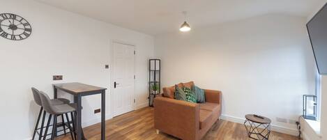 A modern living room with a wooden floor, a brown sofa, a small table with two chairs, a clock on the wall, and a TV on the right.