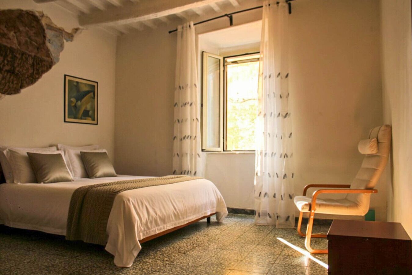 A bedroom with a bed, a chair, a window with curtains, and a painting on the wall.