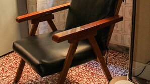 Una silla con estructura de madera y asiento negro, ubicada junto a una ventana que ofrece vistas a un muro de piedra.