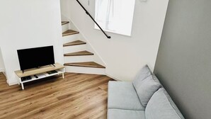 Una sala de estar moderna con un sofá gris, piso de madera, un televisor sobre un mueble bajo y una escalera.