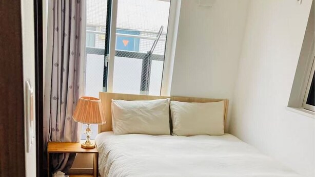 Un dormitorio con cama de madera, una mesita de noche con lámpara y una ventana con vista a edificios.