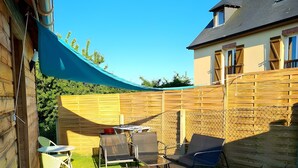 A wooden deck with outdoor furniture, a blue shade sail, and a stone wall.