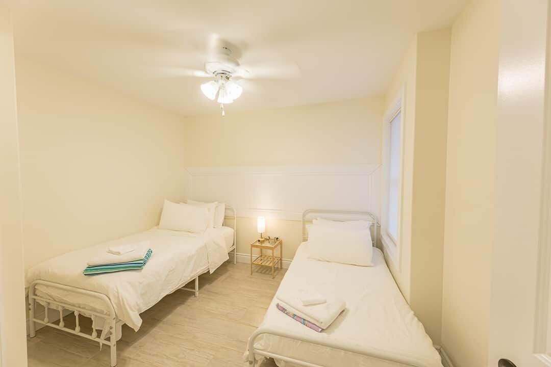 A small, well-lit room with two single beds, a ceiling fan, and a nightstand with a lamp.