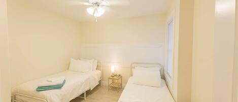 A small, well-lit room with two single beds, a ceiling fan, and a nightstand with a lamp.