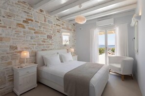 A bedroom with a stone wall, a bed with white bedding, a bedside table with a lamp, and a view of the sea through a balcony door. - Villa Caelia,, Villa (Paros)