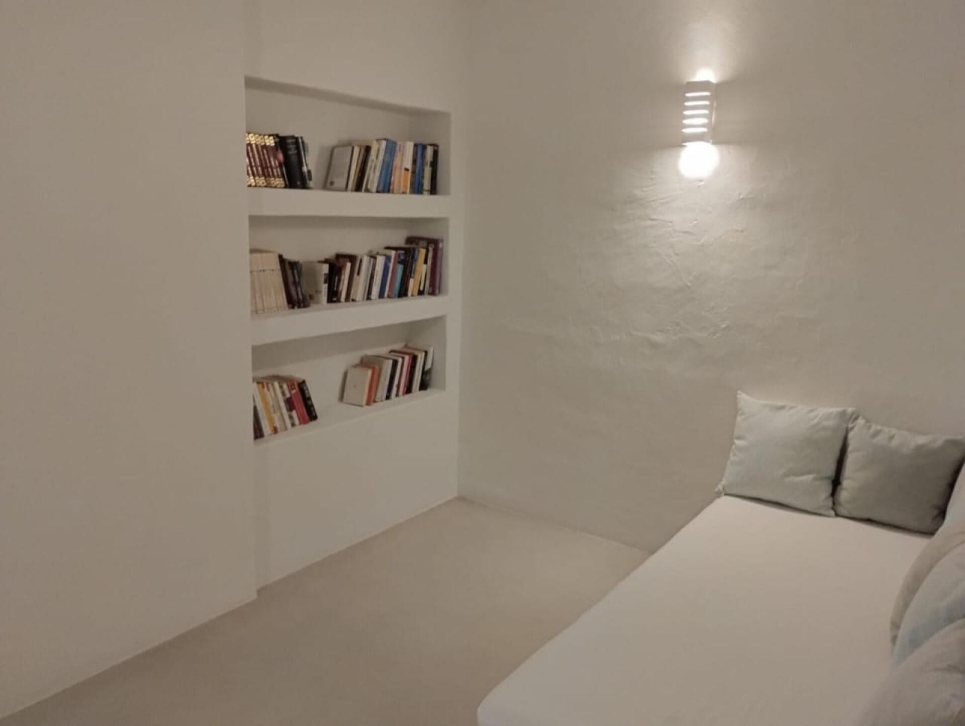 Minimalistinen makuuhuone, jossa on valkoinen sänky, kirjahylly ja seinään kiinnitetty valaisin.