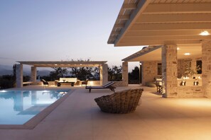 A poolside area with a pergola, lounge chairs, and a stone building with a bar. - Villa Caelia,, Villa (Paros)