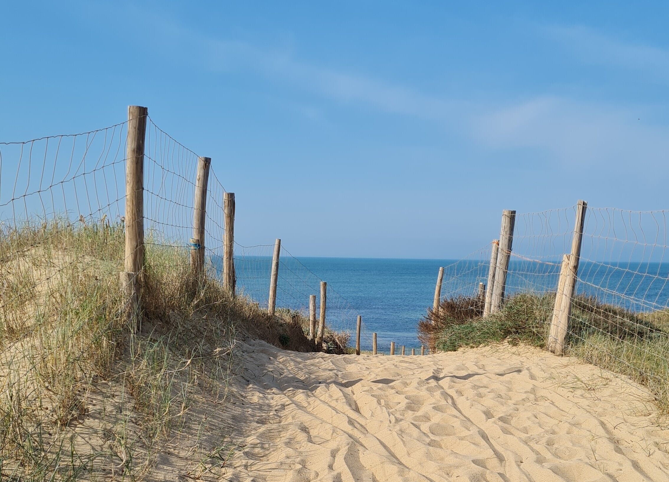 Ein sandiger Strandweg führt zum Meer, umgeben von Holzbefestigungen und Drahtzaun.