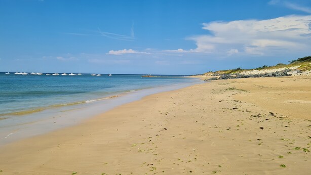 Une plage de sable fin aux eaux calmes, avec des bateaux visibles au loin et une côte rocheuse.
