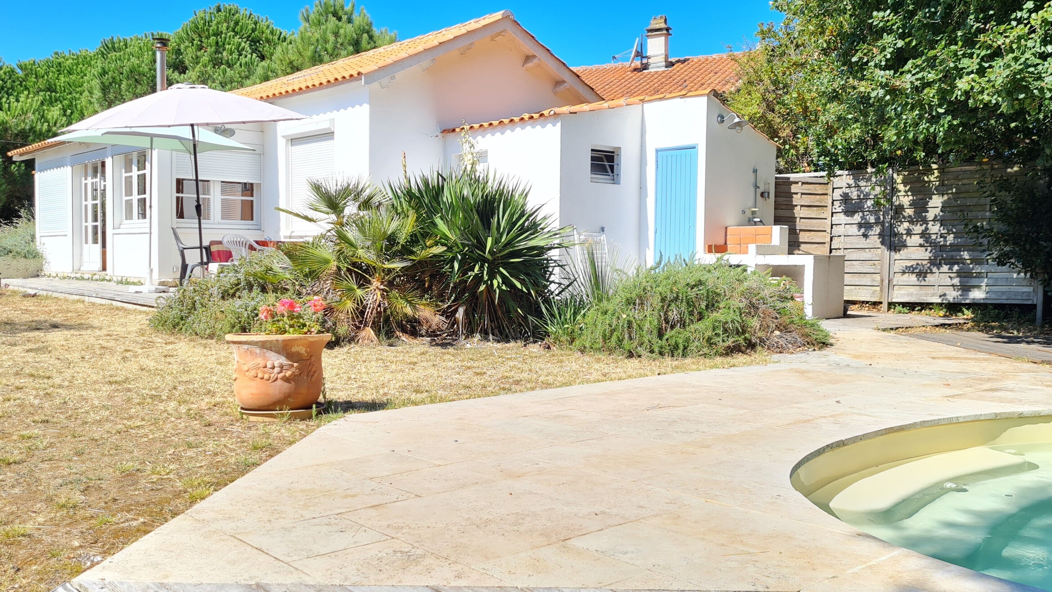 A white house with a tiled roof, a blue door, and a small garden with a patio table and chairs.