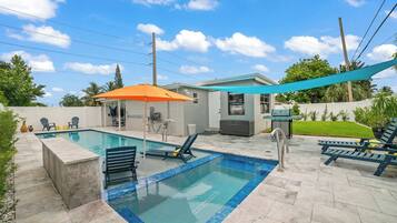 A rectangular swimming pool with a tiled edge, surrounded by lounge chairs and a barbecue grill.