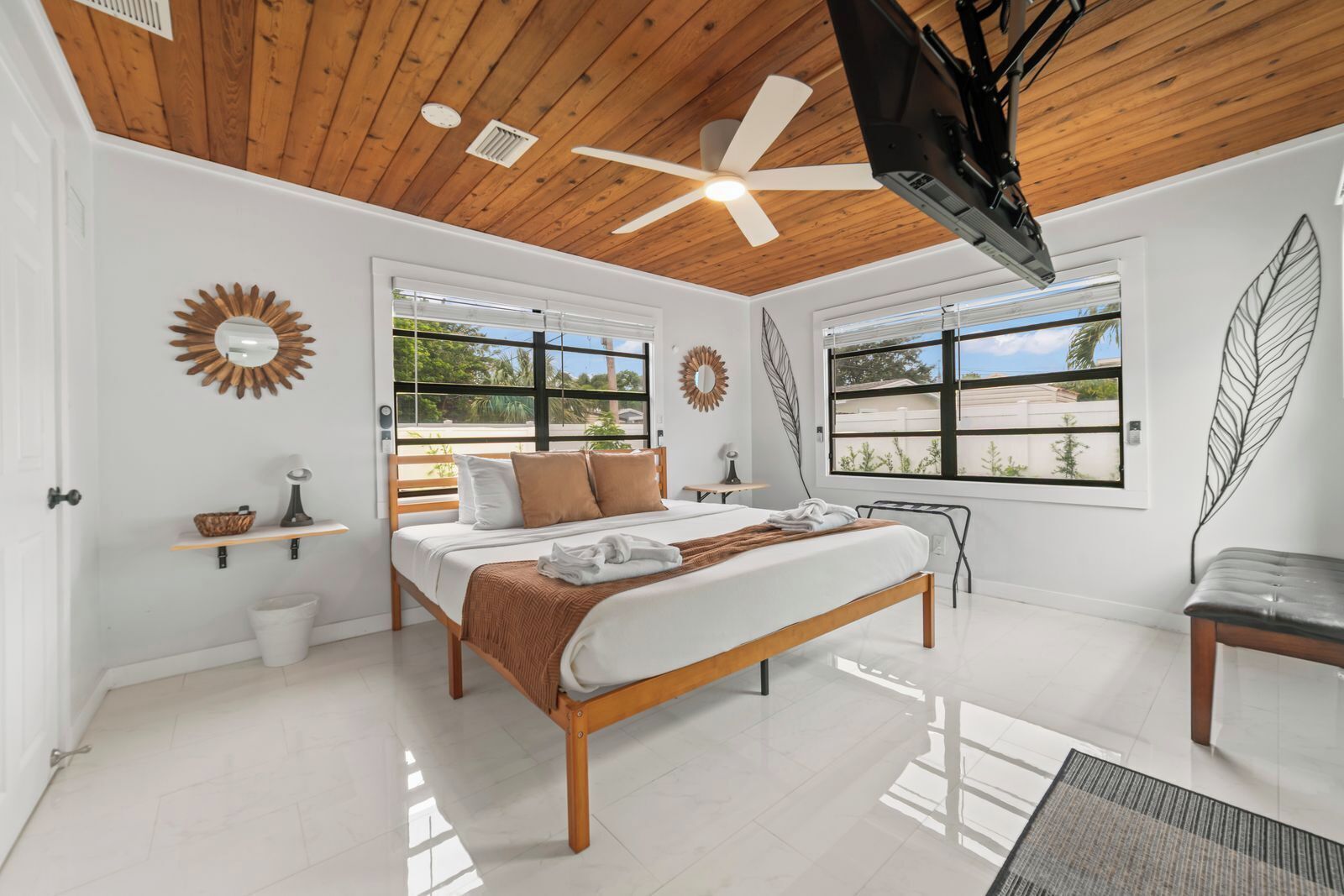 Un dormitorio moderno con techo de madera, cama con colcha blanca y marrón, ventilador de techo y ventanas amplias que ofrecen vistas al verdor.