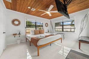 Ein modernes Schlafzimmer mit Holzbalkendecke, einem Bett mit weißer und brauner Bettdecke, einem Deckenventilator und großen Fenstern mit Blick auf Grünflächen.