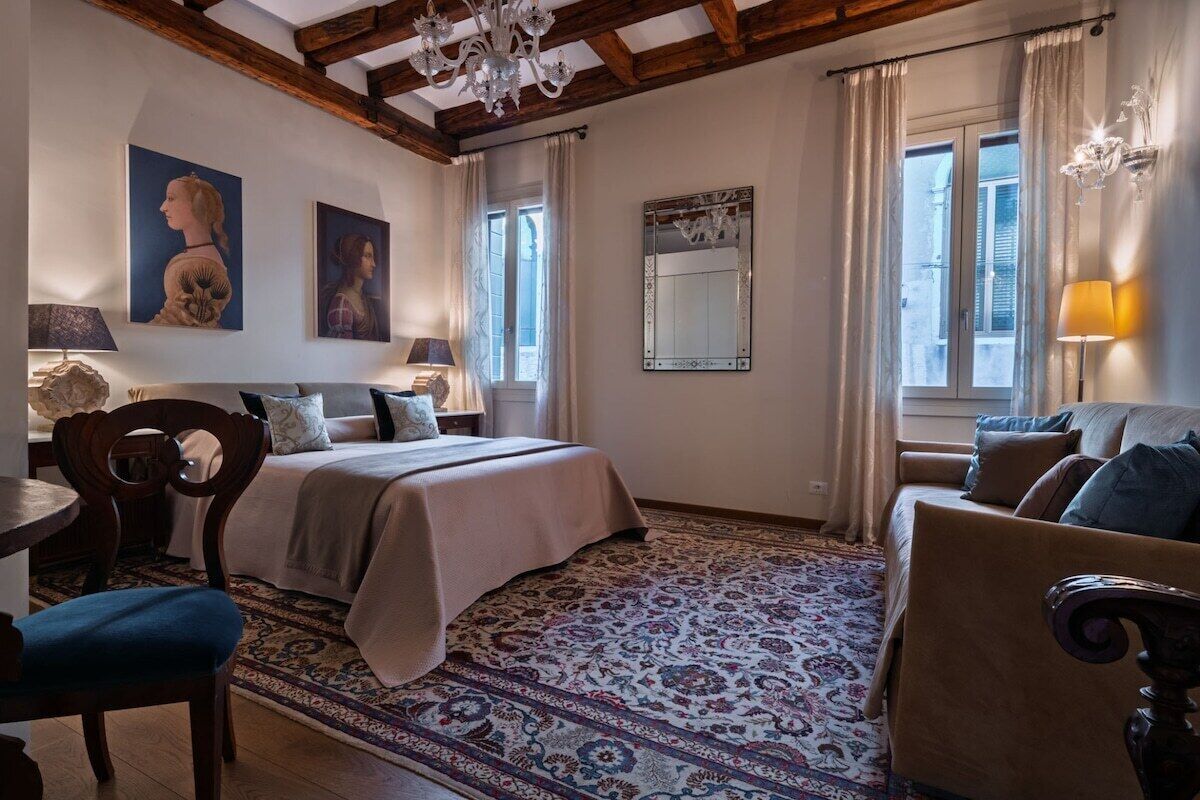 A bedroom with a bed, a sofa, a chair, a desk, a chandelier, and artwork on the walls.