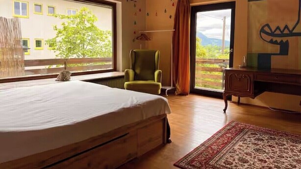 Ein Schlafzimmer mit Bett, Sessel, Schreibtisch und einem großen Fenster mit Blick auf Grünflächen und ein Gebäude.